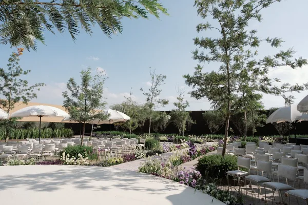 Outdoor ceremony setup with white ceremony chairs and parasols, a curved aisle lined with low flowers and greenery under trees and blue sky