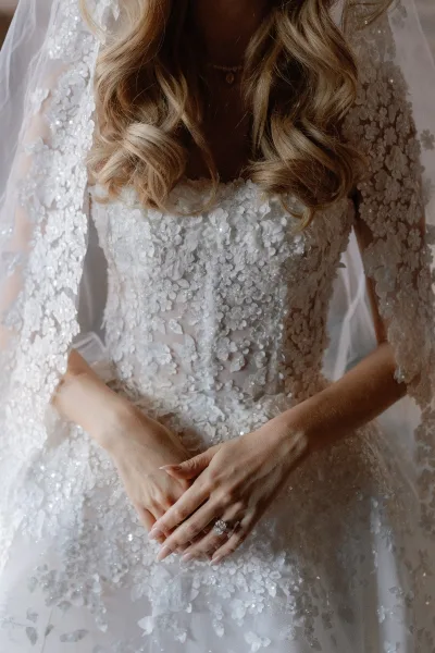 Wedding dress close-up showing lace wedding dress bodice with floral appliqué and sheer sleeves, veil draped, ringed hand in window light