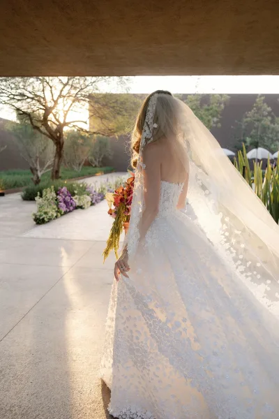 Bridal portrait of a bride from behind holding bouquet, long lace veil flowing over a strapless lace wedding dress in sunset garden patio light