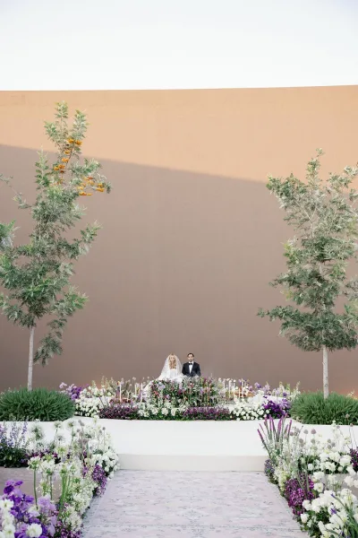 Ceremony setup with modern ceremony backdrop, floral aisle arrangements and candles on a white platform before a stucco wall and trees