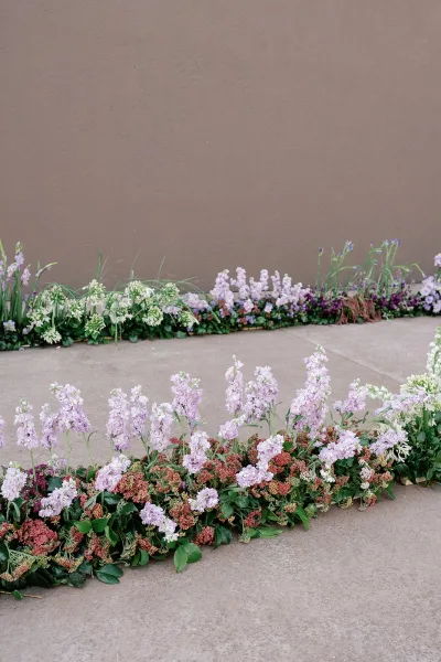 Ceremony aisle flowers in pastel ground arrangements with greenery, accented by purple and white blooms along a concrete walkway by a stucco wall