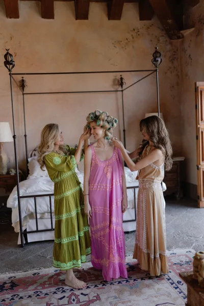 Bridal getting ready as bride in hair rollers wears necklace and earrings with bridesmaids in maxi dresses in a plaster-walled bedroom