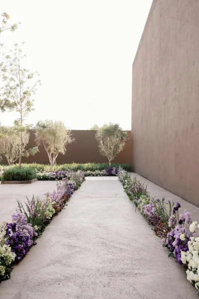 Ceremony aisle design with ground floral aisle of purple, lavender, and white blooms with greenery along a concrete courtyard walkway