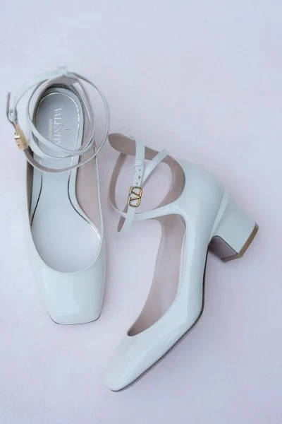 Bridal shoes with ankle straps and gold buckles, glossy white block heels arranged on a soft pink fabric backdrop