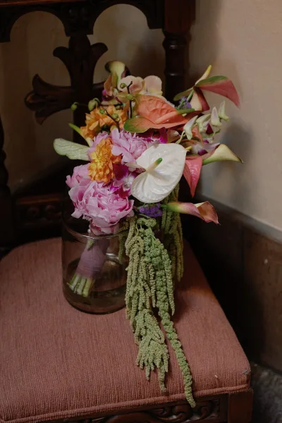 Wedding bouquet with calla lily bridal bouquet blooms in pink and orange, wrapped with ribbon and set in a glass jar on a wooden chair