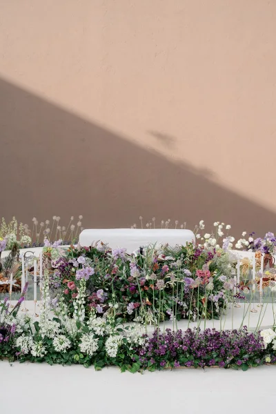 Reception sweetheart table with sweetheart table flowers, lush purple and white blooms, greenery garland, and gold taper candles against a sunlit wall