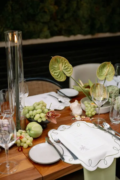 Reception tablescape with wedding table setting of green grapes, apples, garlic, anthurium and orchids, taper candle in glass hurricane, greenery wall backdrop