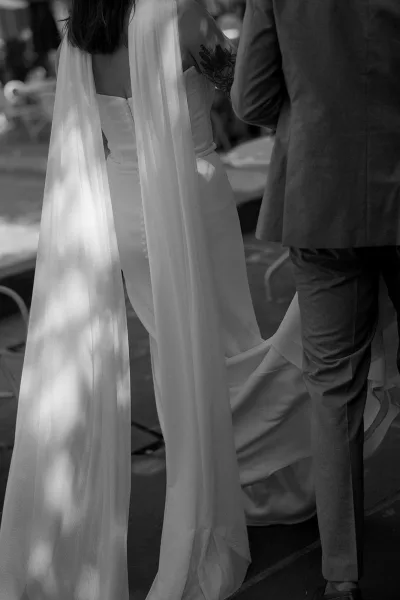 Ceremony moment in a black and white wedding photo, bride and groom from behind with veil train in dappled sunlight by trees