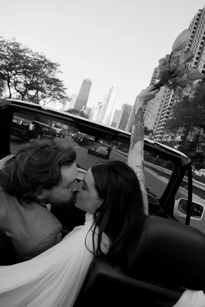 Wedding kiss portrait of bride and groom kissing in a convertible, bride lifting bouquet, city skyline and traffic behind
