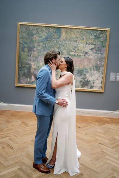 Wedding kiss portrait of bride and groom kissing, her cape veil and white gown against an art gallery wall with framed artwork and placards