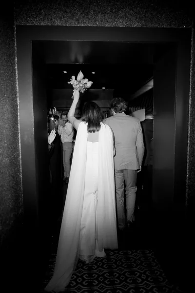 Wedding reception entrance as bride in a bridal cape raises her bouquet overhead beside groom in a light suit, entering doorway past cheering guests