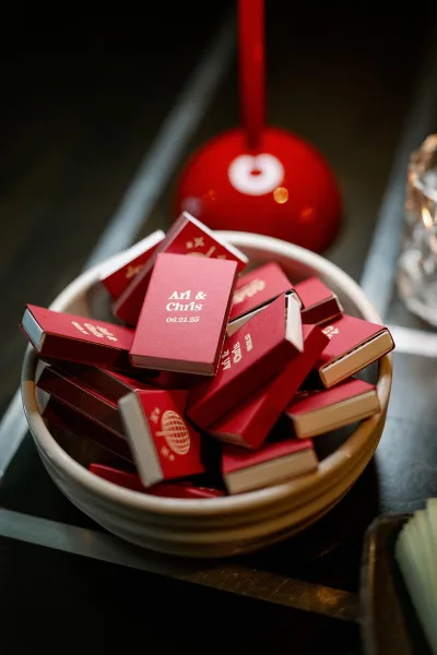 Wedding matchboxes and custom wedding matchboxes piled in a ceramic bowl on a dark tabletop with glassware and a red accent nearby