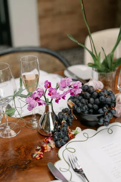 Reception tablescape on a wood wedding table with purple orchid bud vases, grapes, garlic, scalloped menu cards, and flutes with plates and flatware