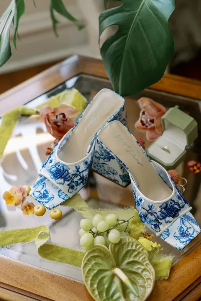 Bridal shoes, blue and white bridal heels styled on a glass tabletop with velvet ribbon, ring box, orchids, anthurium, and green grapes