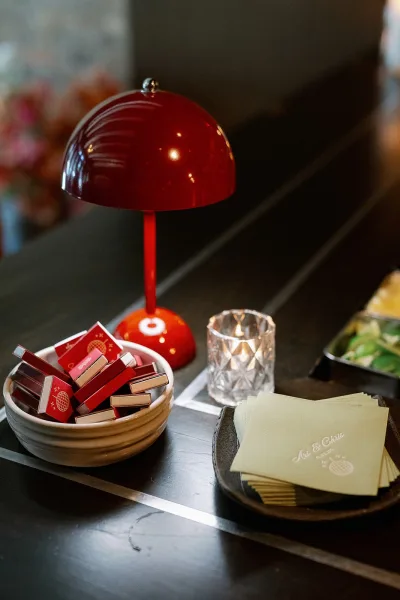 Wedding matchbooks and custom wedding matchbooks arranged on a black tray beside a red table lamp, crystal votive, and ceramic bowl on dark tabletop
