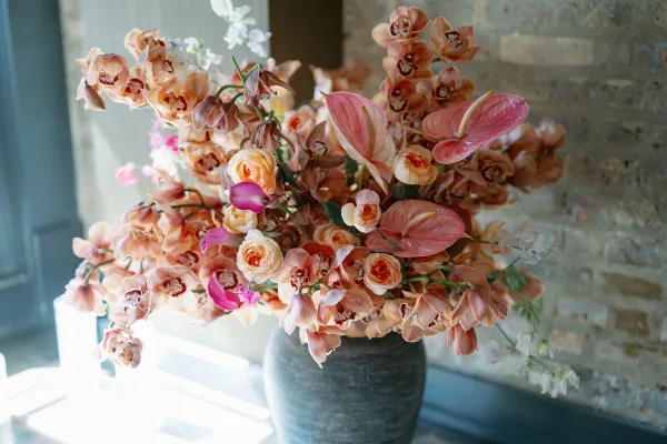 Wedding floral arrangement with orchids, anthurium, and garden roses in a ceramic vase, softly lit by a window against a brick wall