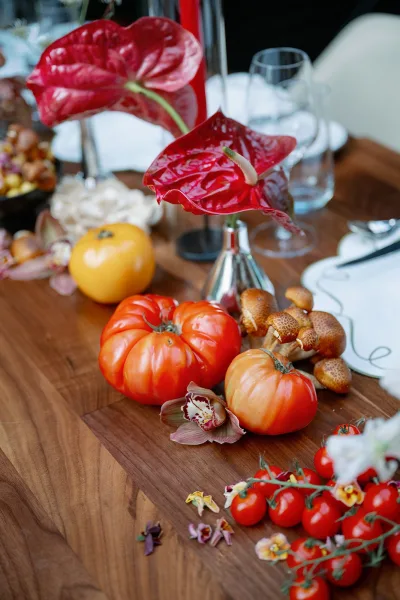 Reception tablescape with tomato wedding centerpiece featuring red anthurium, heirloom tomatoes, citrus, mushrooms, and glassware on a wood table