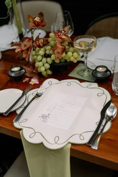 Reception tablescape with a wedding place setting featuring scalloped charger, menu card, silver flatware, orchids, grapes, and candlelight on a wood table