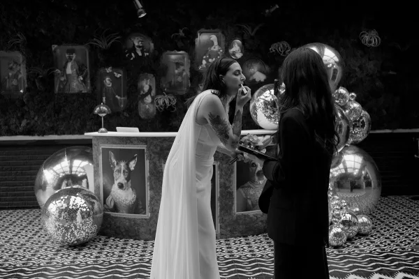 Bridal makeup touch-up as bride applies lipstick in veil and gown, holding a handheld mirror near a bar counter with disco balls backdrop