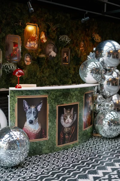 Wedding photo booth with a moss wall photo booth backdrop, framed pet portraits, and disco ball balloons beside a bar counter under track lights
