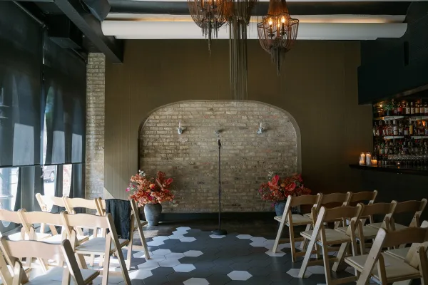 Indoor ceremony setup with wood folding chairs and statement urn florals facing a brick arch wall, chandelier overhead and candlelit bar shelves nearby