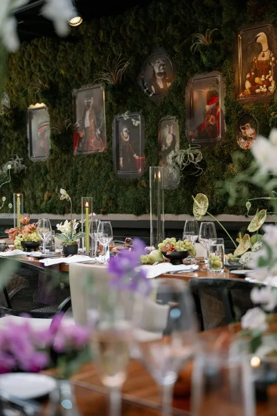 Reception tablescape with wood table reception decor, white plates, wine glasses, cylinder and taper candles, orchids and grapes before a moss wall backdrop
