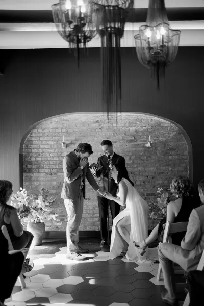 Wedding vows during an indoor wedding ceremony as bride and groom hold hands by a microphone, with brick wall alcove and chandeliers behind.