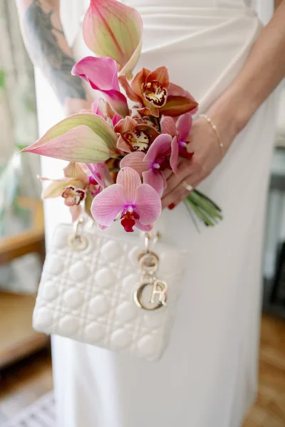 Bridal bouquet with orchid wedding bouquet blooms and anthurium held by bride in gown, lit by window light on a wooden floor