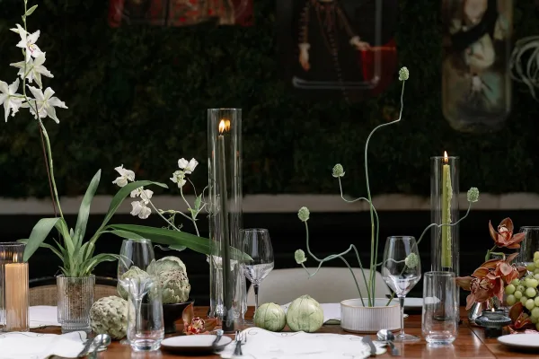 Reception tablescape with a candlelit wedding table of taper candles, orchids, grapes, and artichokes on white linens against dark greenery backdrop
