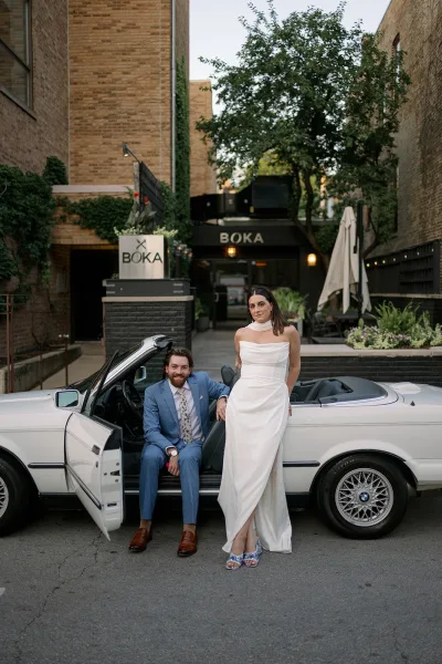 Couple portrait of bride in a strapless wedding dress leaning on a white convertible car beside groom in a blue suit on a city street outside a restaurant entrance