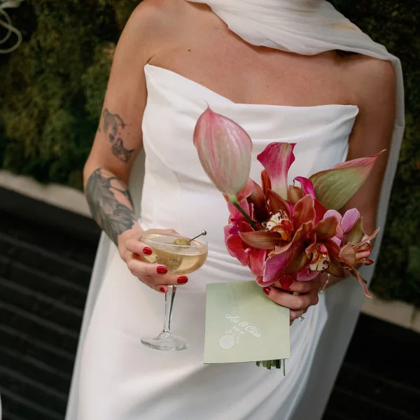Bridal portrait of a tattooed bride in a strapless wedding dress holding an orchid bouquet with menu card, cocktail, and red nails by hedges