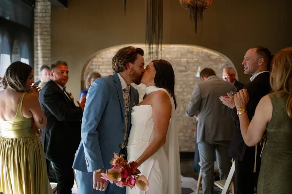 Wedding kiss as bride in a strapless gown and veil holds a bouquet, kissing groom in a blue suit under a brick archway with guests behind