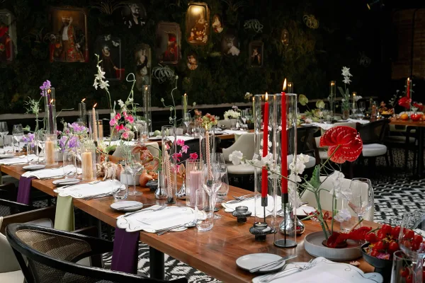 Reception tablescape with long banquet table decor on a wood dining table, white plates, glass cylinders with twisted taper candles, and anthurium florals before a greenery wall