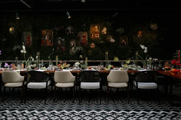 Reception tablescape on a long banquet table with white runner, taper candles, orchids, anthuriums, grapes, and mixed chairs before a moss wall backdrop
