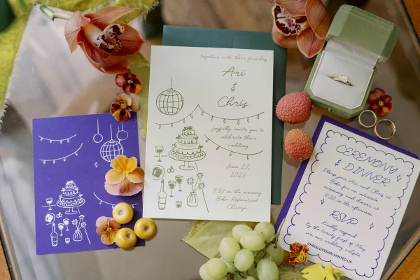 Wedding invitation suite with an illustrated wedding invitation, RSVP card and envelopes, accented by rings, orchids, pansies, fruit and ribbon on a tabletop