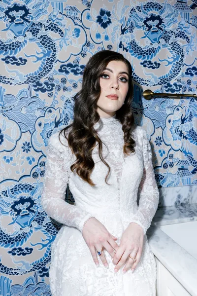 Bridal portrait in a high neck wedding dress with long sleeve lace cuffs, statement ring and earrings, set against blue patterned wallpaper indoors