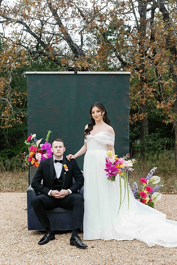 Couple portrait of bride in off the shoulder gown with a bright bouquet beside groom in black tuxedo, set against autumn trees and greenery