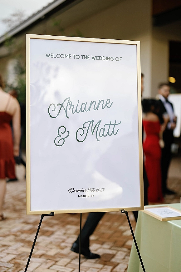 Wedding welcome sign in a gold frame with calligraphy text on an easel, set on a brick patio walkway as guests arrive nearby