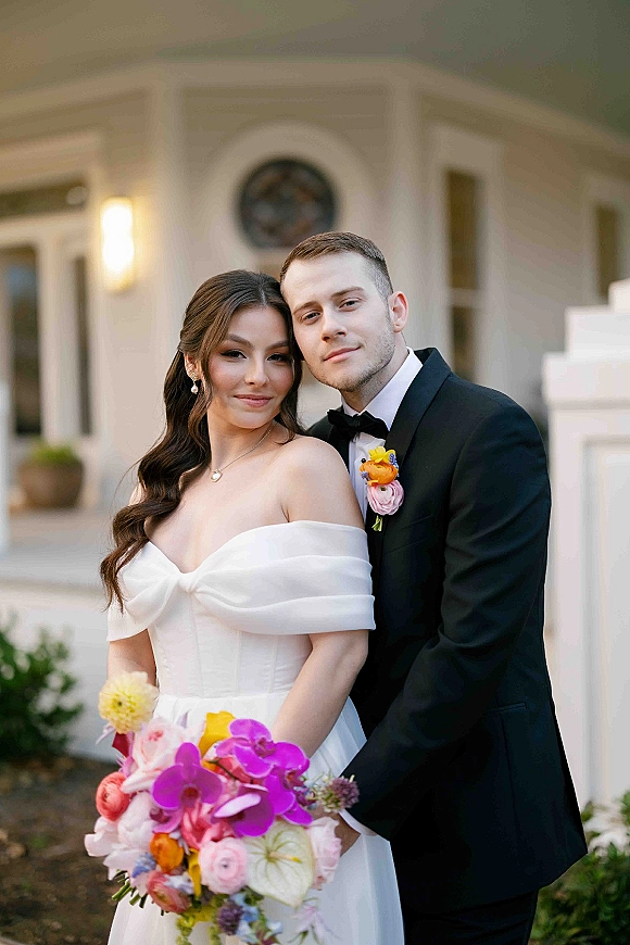 Couple portrait of newlyweds embracing, bride holding a pink and purple bouquet beside a white venue doorway with greenery backdrop