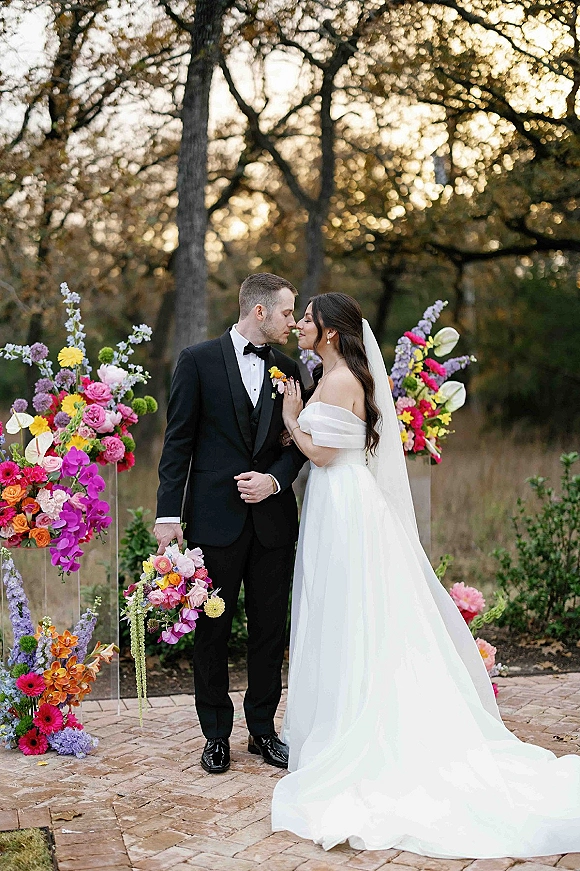 Wedding couple portrait of bride and groom kiss, her off-the-shoulder dress and long veil beside colorful florals on clear acrylic pedestals outdoors