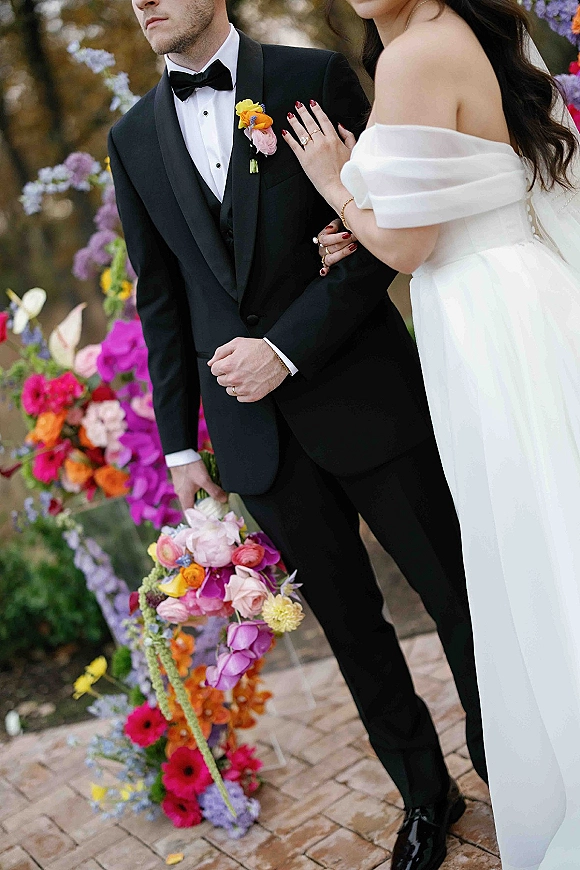 Couple portrait of bride and groom close up, her hand on his black tuxedo showing engagement ring beside a colorful bouquet on a garden walkway