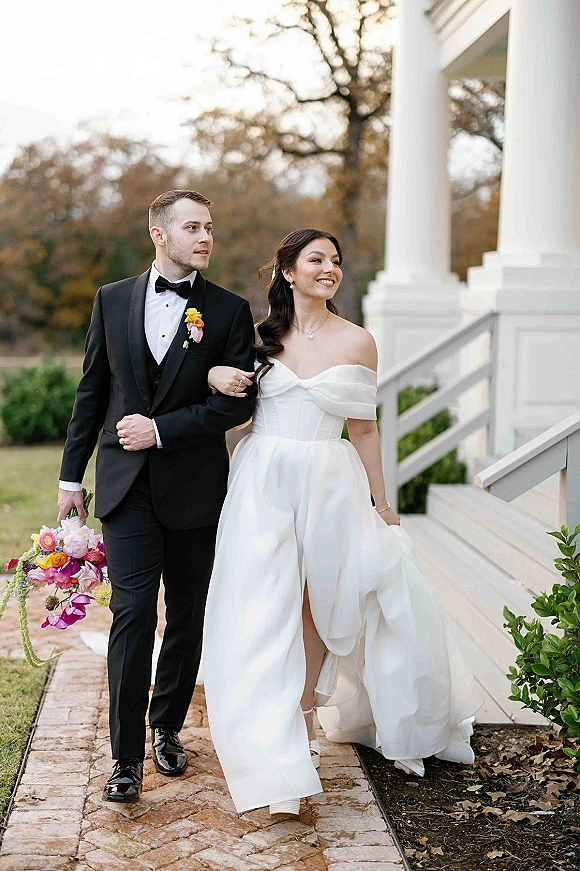 Couple portrait of bride and groom walking arm in arm, groom holding bouquet, by white porch columns on an outdoor walkway with fallen leaves