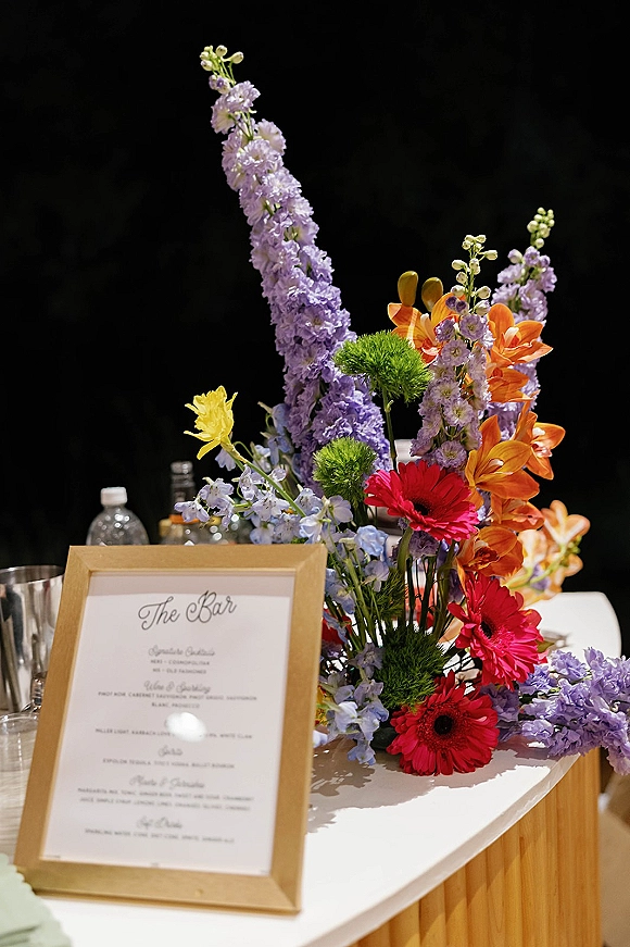 Wedding bar sign in a gold frame displaying a signature drinks sign beside bright florals and glassware on a bar with bottles and black drape