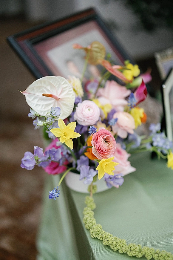 Wedding flowers in a colorful wedding centerpiece with white anthurium, ranunculus, and daffodils beside a framed photo on a tablecloth