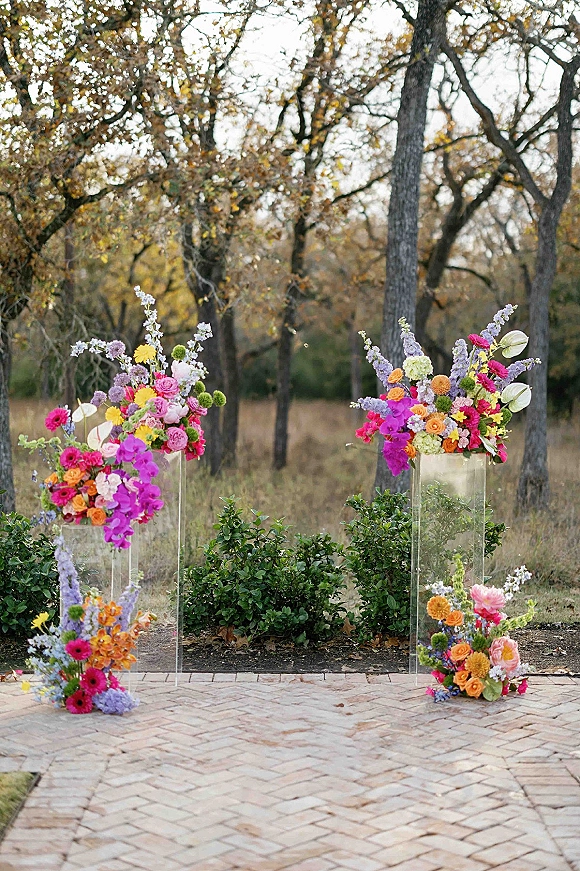 Ceremony floral decor with acrylic pedestal wedding flowers, bright pink and orange arrangements on a brick patio beside trees and grass field