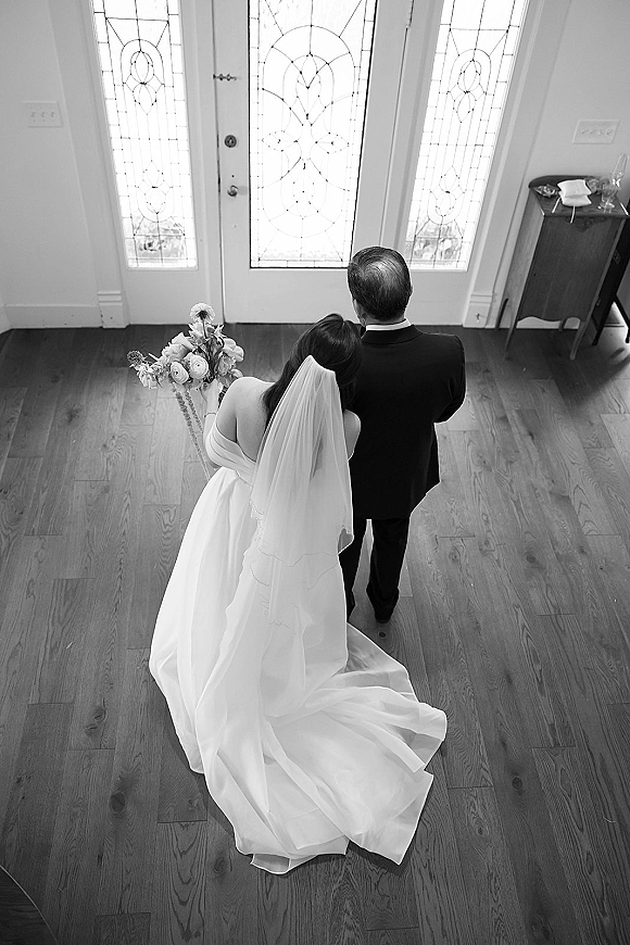 Wedding couple portrait in black and white, overhead view from behind as bride leans on groom, holding bouquet with long veil on wood floor by glass doors