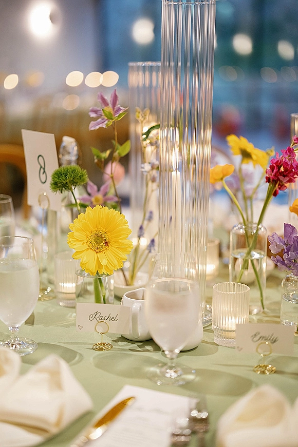 Reception tablescape with colorful wedding tablescape florals in bud vases and tall glass cylinder centerpiece, votive candles, and place cards under blurred lights