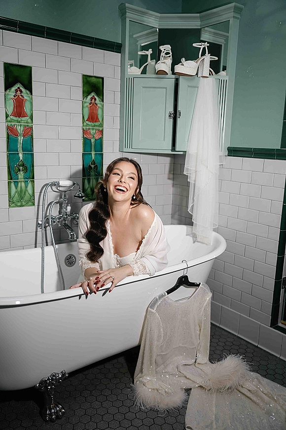 Bridal portrait of a bride in a sequined gown beside a clawfoot bathtub, wearing drop earrings and red manicure in a vintage tiled bathroom