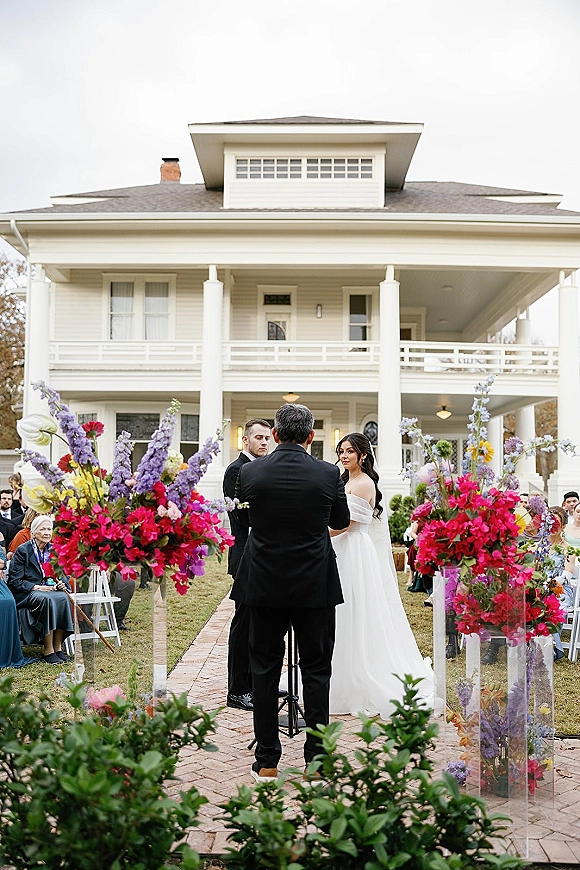 Wedding ceremony with bride and groom exchanging vows beside bright pink and purple florals on clear stands outside a white house porch