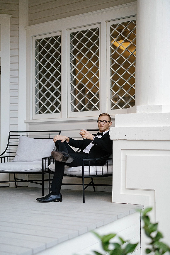 Groom portrait of a tuxedo groom in a black tuxedo and bow tie, wearing eyeglasses and holding a whiskey glass on a white porch bench